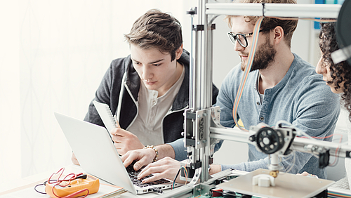 Neues verbinden: An der Fakultät Ingenieurwissenschaften der Hochschule Mittweida starten im Herbst 2026 zwei neue Studiengänge. Zwei junge Männer sitzen vor einem Laptop. Neben ihnen steht ein 3D-Drucker, der ein Plastikteil herstellt.